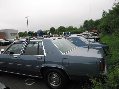 1991 Ford LTD Crown Victoria - Side-rear view of a blue Ford LTD Crown Victoria police sedan with roof spotlight, parked in a lot with other cars.