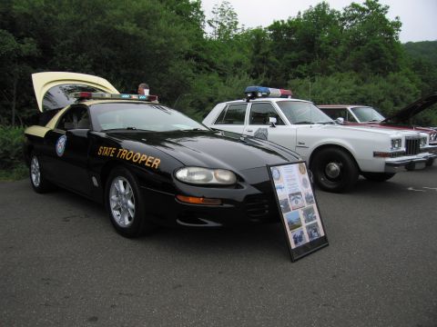 1991 Ford LTD Crown Victoria - black-and-tan Chevrolet Camaro marked 'STATE TROOPER' with its hood up and an informational placard beside it.