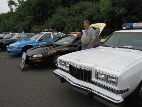 1991 Ford LTD Crown Victoria - man stands by an open hood among a lineup of vintage police cars including a black State Trooper coupe and a white sedan.