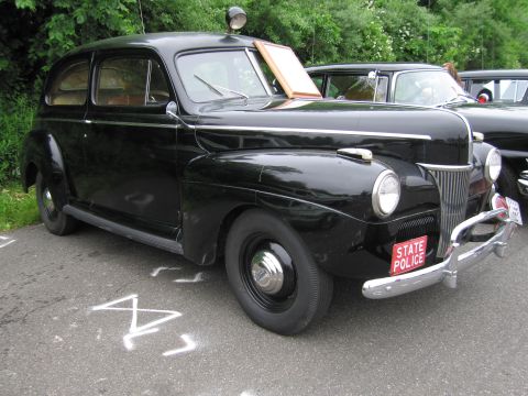 1991 Ford LTD Crown Victoria - restored black 1940s two-door sedan with a roof spotlight and a red STATE POLICE plate parked on asphalt.