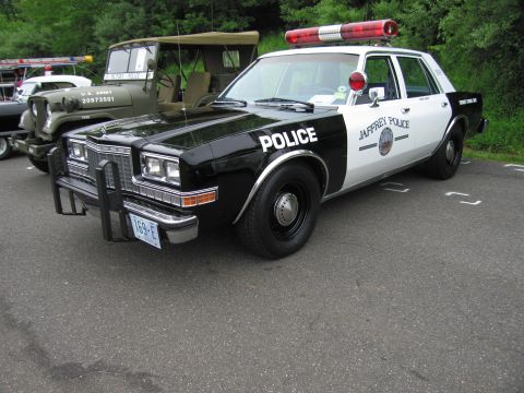 1991 Ford LTD Crown Victoria - black-and-white Jaffrey Police sedan with a chrome push bar and roof light bar parked beside a military vehicle.