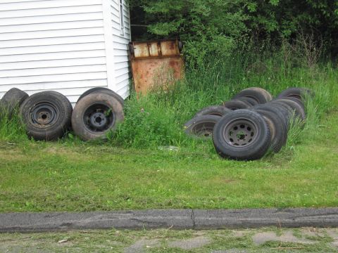 1991 Ford LTD Crown Victoria - Several worn tires and steel wheels lie in the grass beside a white-sided building and a rusty metal panel.