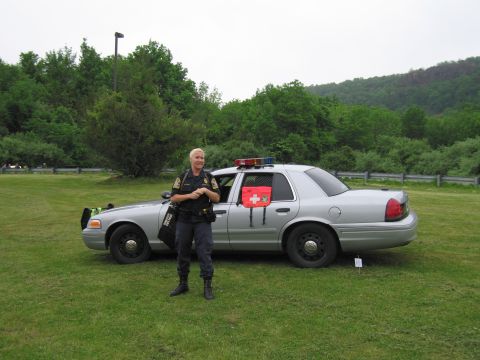 1991 Ford LTD Crown Victoria - uniformed officer stands beside a silver State Police Crown Victoria with a roof light bar on a grassy field.
