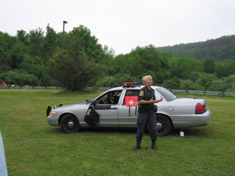 1991 Ford LTD Crown Victoria - blonde uniformed officer gestures while speaking beside a silver State Police sedan on a grassy field.