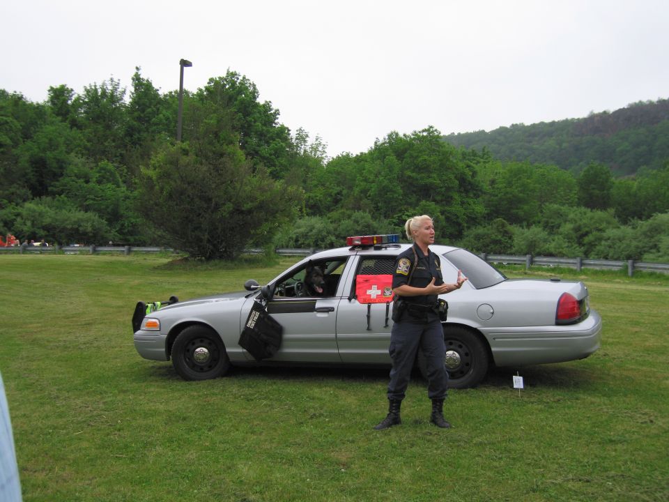 Blonde officer speaking beside a silver State Police sedan on grass