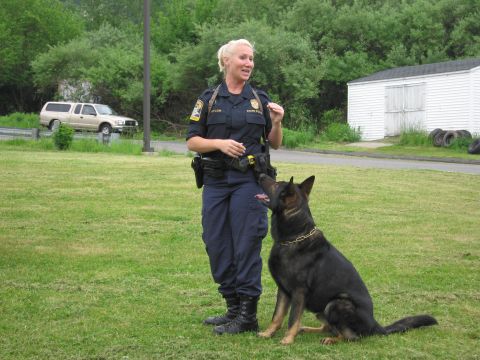 1991 Ford LTD Crown Victoria - blonde uniformed officer smiles while her German Shepherd K9 sits looking up at her on a grassy field.