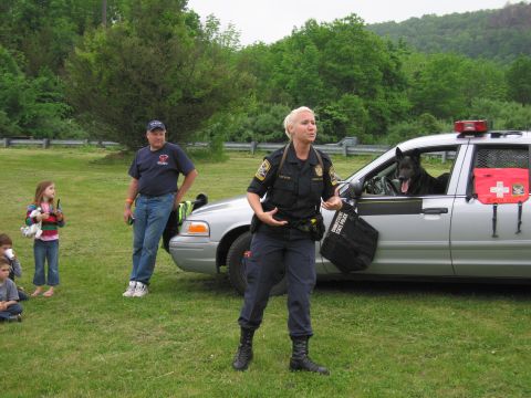 1991 Ford LTD Crown Victoria - blonde female officer in uniform speaks and gestures in front of a silver police cruiser with a dark dog visible in the rear window, specta…