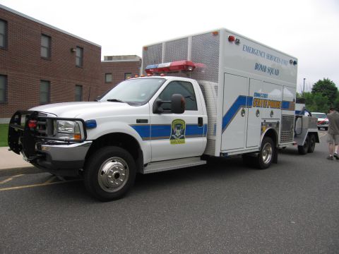 1991 Ford LTD Crown Victoria - white Ford State Police Bomb Squad utility truck with blue striping tows a trailer in a parking lot beside a brick building, a person stand…