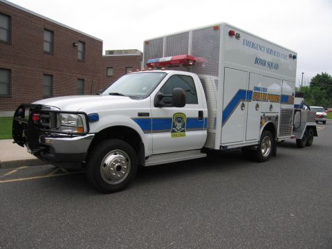 1991 Ford LTD Crown Victoria - white Ford State Police Emergency Services Unit Bomb Squad box truck with blue stripes parked at an angle, towing a trailer near a brick bu…