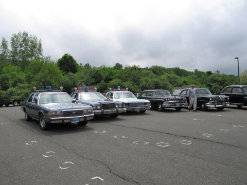 1991 Ford LTD Crown Victoria - lineup of classic police cars in a paved lot, including blue Ford sedans with light bars and older black sedans, under an overcast sky.