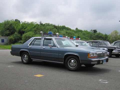 1991 Ford LTD Crown Victoria - blue Ford LTD Crown Victoria State Police sedan with a roof light bar parked in a lot, with other classic cars behind it under a gray sky.