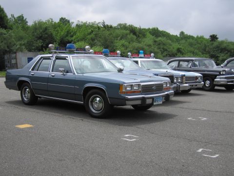 1991 Ford LTD Crown Victoria - blue Ford LTD Crown Victoria State Police cruiser leads an angled row of classic police cars with State Police light bars in a parking lot.