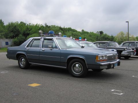 1991 Ford LTD Crown Victoria - blue Ford LTD Crown Victoria State Police sedan with a roof light bar sits in a paved lot with other classic cars under an overcast sky.