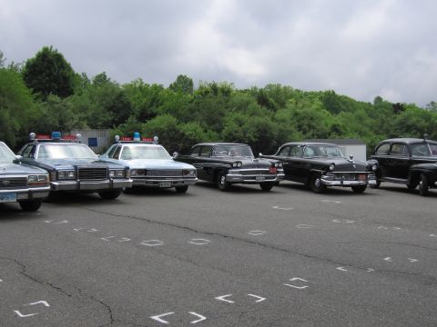 1991 Ford LTD Crown Victoria - angled lineup of classic police cars in a paved lot, mixing blue and black sedans with roof beacons, under a gray overcast sky.