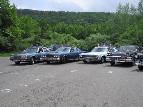 1991 Ford LTD Crown Victoria - Four classic police sedans, two blue Fords and two darker cars with roof beacons, parked side by side in a lot edged by green trees.