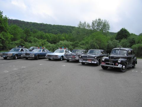 1991 Ford LTD Crown Victoria - row of six classic police cars in a lot, progressing from a blue Ford Crown Victoria to a black 1940s-style sedan, against a wooded hillsid…