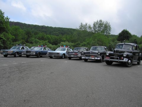 1991 Ford LTD Crown Victoria - angled lineup of six classic police cars on pavement, ending in a black 1940s-style Ford sedan, with a green wooded hillside behind.
