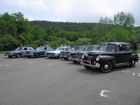 1991 Ford LTD Crown Victoria - black 1940s-style Ford police sedan sits at the front of an angled row of classic police cars in a paved lot under a cloudy sky.