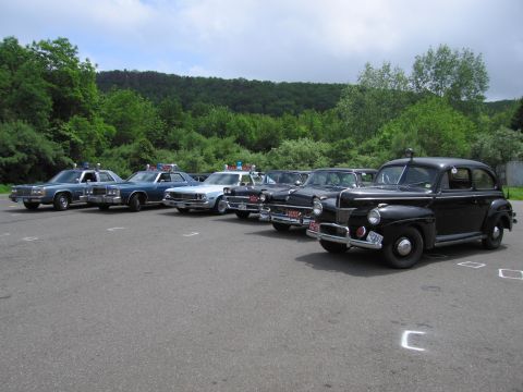 1991 Ford LTD Crown Victoria - black 1940s-style Ford police sedan leads an angled lineup of classic police cars across a paved lot bordered by green trees.