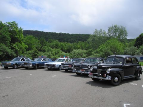 1991 Ford LTD Crown Victoria - black 1940s-style Ford police sedan anchors an angled row of classic police cruisers on pavement, with a wooded ridge in the background.