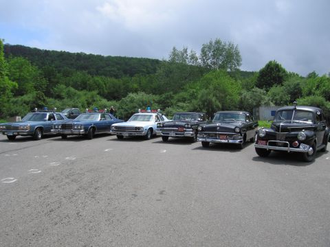 1991 Ford LTD Crown Victoria - Six classic police cars line a paved lot, ranging from a blue Ford Crown Victoria to a black 1940s-style sedan, beneath a wooded hillside.