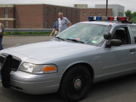 1991 Ford LTD Crown Victoria - Front three-quarter view of a silver Ford Crown Victoria police cruiser with a roof light bar, two men standing near it by a brick building.