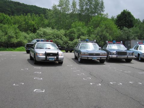 1991 Ford LTD Crown Victoria - silver Ford Crown Victoria police cruiser sits beside a row of older blue Ford police sedans with roof beacons in a paved lot edged by tree…
