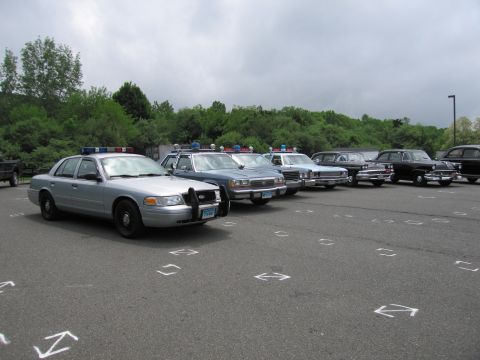 1991 Ford LTD Crown Victoria - diagonal row of police cars from a silver Crown Victoria to older sedans, parked in an arrow-marked lot under cloudy skies.