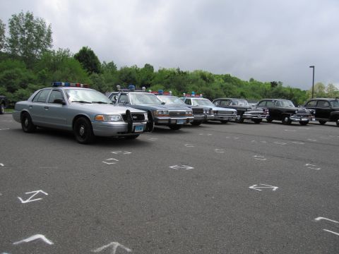 1991 Ford LTD Crown Victoria - lineup of police cruisers led by a silver Ford sedan, parked at an angle in a marked lot beside green trees.