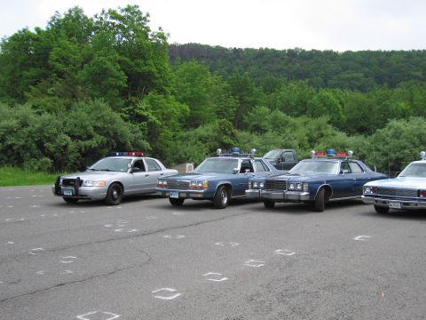 1991 Ford LTD Crown Victoria - silver Ford police interceptor parked beside older blue police sedans in a lot backed by green wooded hills.