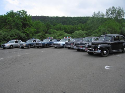 1991 Ford LTD Crown Victoria - long diagonal row of police cars spanning a modern silver cruiser to a vintage black coupe, parked before wooded hills.