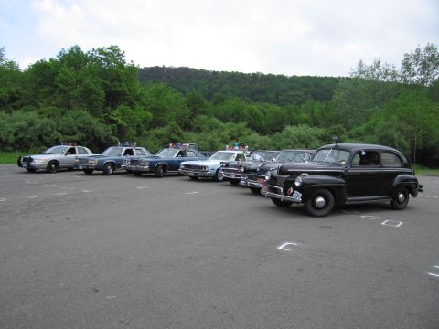1991 Ford LTD Crown Victoria - row of police cruisers led at front by a black vintage sedan, lined up in a marked lot before green hills.