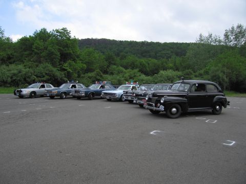 1991 Ford LTD Crown Victoria - lineup of police cars with a black vintage sedan at right and a silver modern cruiser at left, parked before wooded hills.