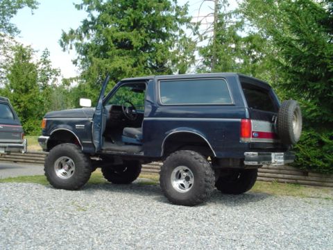 1990 Ford Bronco - A lifted 1987 Ford Bronco with its driver's door open sits parked on a gravel lot.