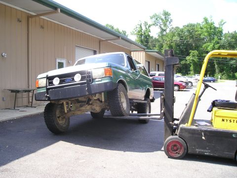 1989 Ford Bronco - Forklift lifts a green Ford Bronco, showing its front end and suspension.