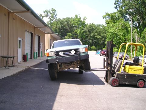 1989 Ford Bronco - A green Ford Bronco is being lifted by a forklift, with other vehicles parked in the background.