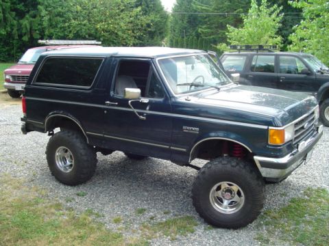1990 Ford Bronco - This lifted 1990 Ford Bronco features a dark blue paint job and aftermarket wheels, parked outdoors with other vehicles visible.