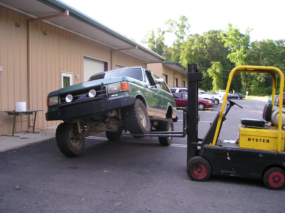 Green Ford Bronco hoisted in the air on a yellow Hyster forklift outside a metal shop