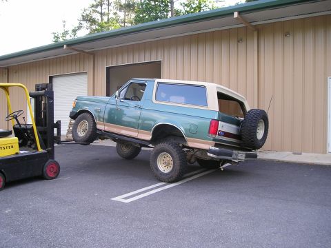 1989 Ford Bronco - Green and tan full-size Ford Bronco propped nose-up on a wooden pallet by a yellow Hyster forklift outside a tan metal shop building, expos…