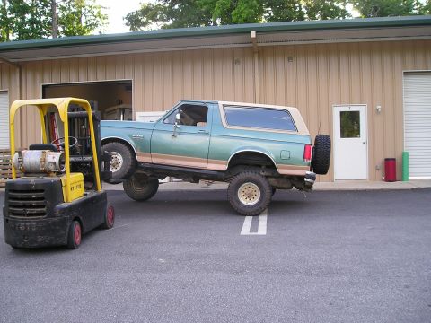 1989 Ford Bronco - A green and tan Ford Bronco is lifted by a yellow forklift in front of a tan building.