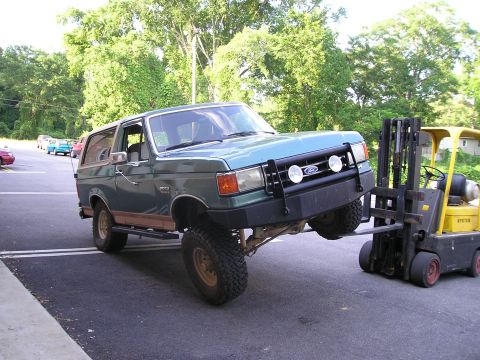 1989 Ford Bronco - Green full-size Ford Bronco with tan hardtop and lower two-tone lifted at the front by a yellow Hyster forklift under its front bumper, lea…