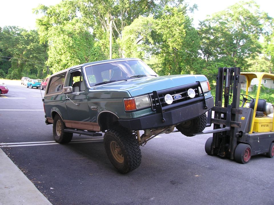 Green Ford Bronco lifted at the front by a yellow Hyster forklift in a parking lot