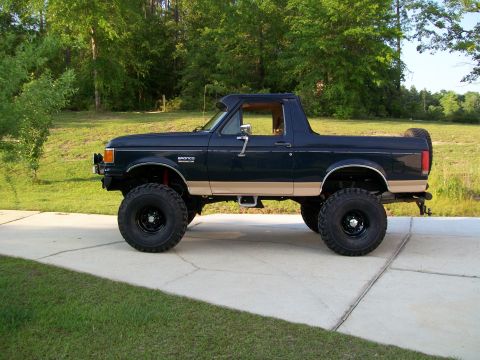1990 Ford Bronco - This lifted Ford Bronco is parked on a concrete driveway with a grassy area and trees in the background.
