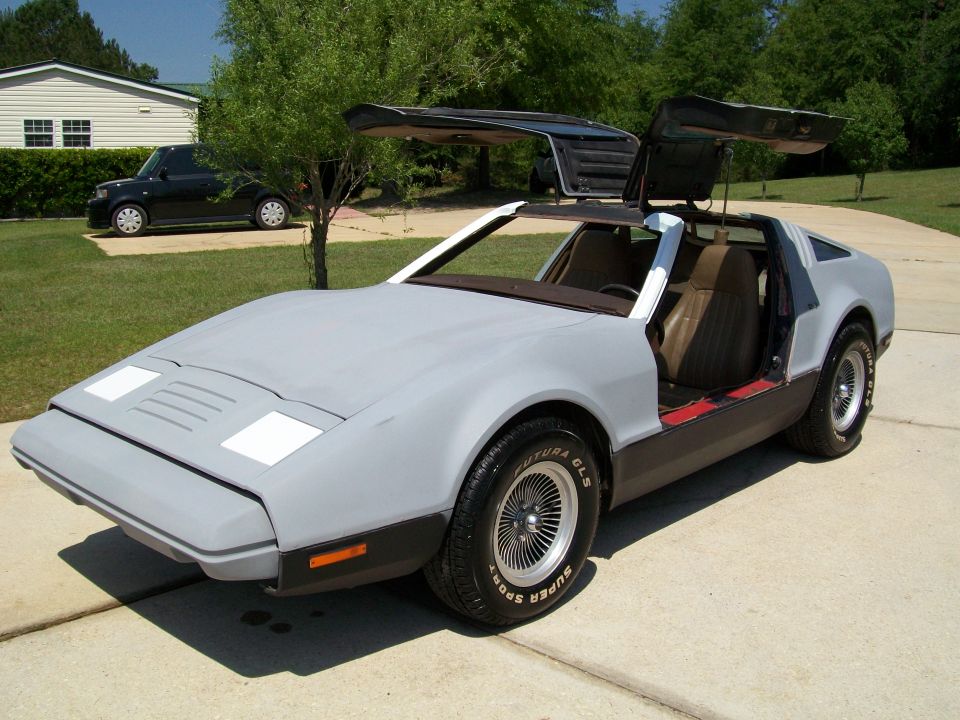 Gray gullwing-door sports coupe with doors raised, parked on a concrete driveway, front three-quarter view