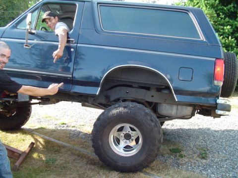 1990 Ford Bronco - A dark blue two-door 1990 Ford Bronco SUV lifted on a suspension/axle swap, shown from the passenger side on a gravel driveway with oversiz…