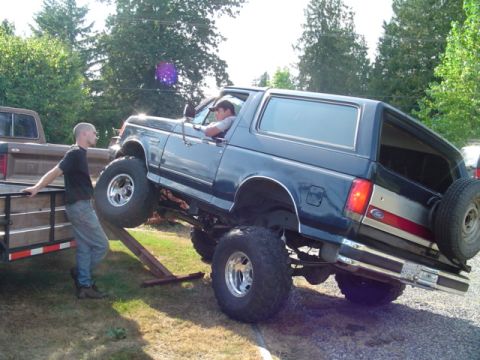 1990 Ford Bronco - Lifted dark-blue Ford Bronco full-size SUV climbing wooden ramps onto a utility trailer hitched to a brown pickup, front wheels already up…