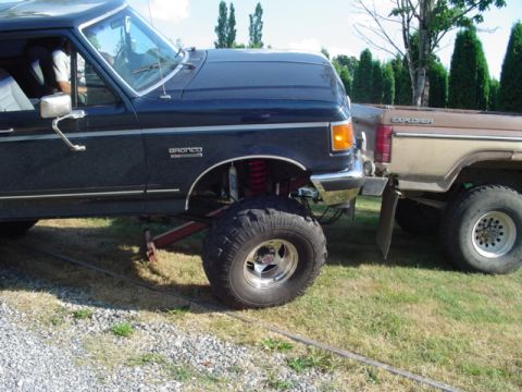 1990 Ford Bronco - Side view of the front half of a lifted dark blue Ford Bronco with a solid front axle, red coil springs, red-painted steering and radius-ar…