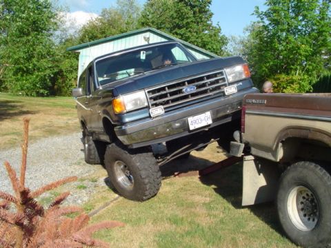 1990 Ford Bronco - Dark blue late-1980s Ford Bronco with a tall suspension lift and oversized tires on a gravel driveway, its front end raised high in the air…