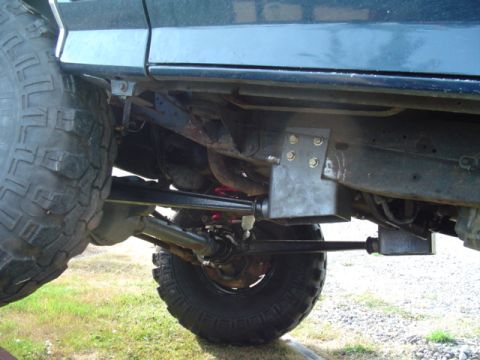 1990 Ford Bronco - Low view under the front of a dark-colored lifted 4x4 showing a solid front axle with crossover steering, drag link and tie rod, and freshl…