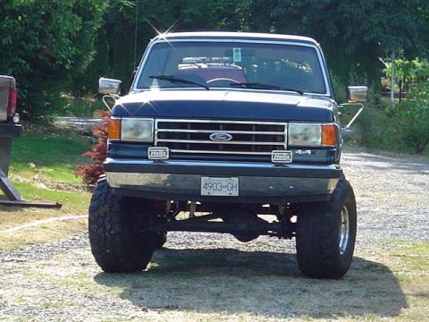 1990 Ford Bronco - Straight-on front view of a lifted dark-blue Ford Bronco parked on a gravel driveway, showing the chrome grille with Ford oval, a 4x4 badge…
